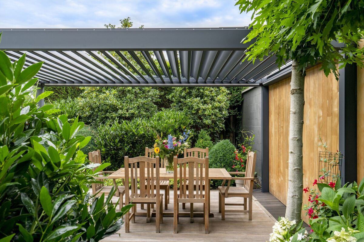Pergola mit Lamellendach über einer Holzterrasse mit Esstisch, umgeben von Pflanzen